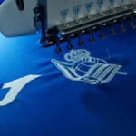 Máquina bordando el escudo de la Real Sociedad junto al logo de Joma en una camiseta azul del club txuri-urdin