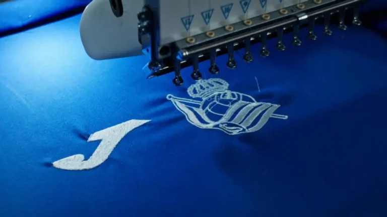 Máquina bordando el escudo de la Real Sociedad junto al logo de Joma en una camiseta azul del club txuri-urdin