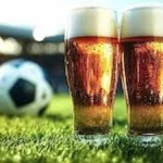 Dos vasos de cerveza con espuma dorada sobre el césped de un estadio, con un balón de fútbol desenfocado al fondo