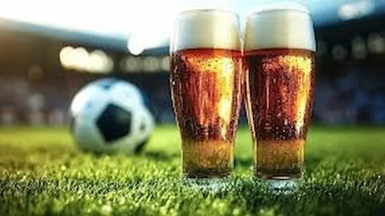 Dos vasos de cerveza con espuma dorada sobre el césped de un estadio, con un balón de fútbol desenfocado al fondo