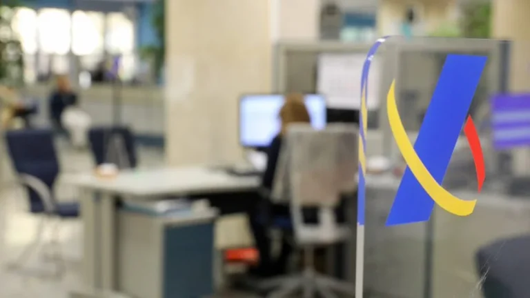 Anagrama de la Agencia Tributaria sobre cristal en una oficina de Hacienda con una empleada trabajando al fondo
