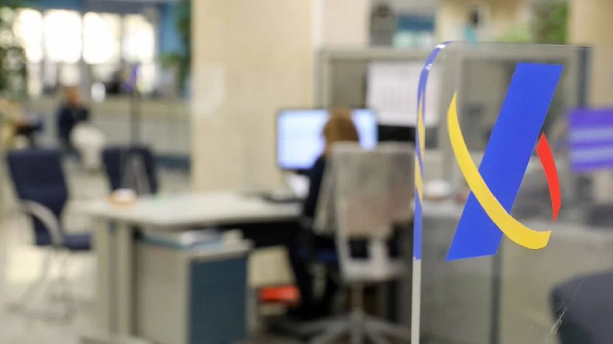 Anagrama de la Agencia Tributaria sobre cristal en una oficina de Hacienda con una empleada trabajando al fondo