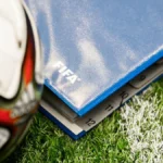 Balón de fútbol junto a una carpeta azul con el logo de la FIFA sobre el césped