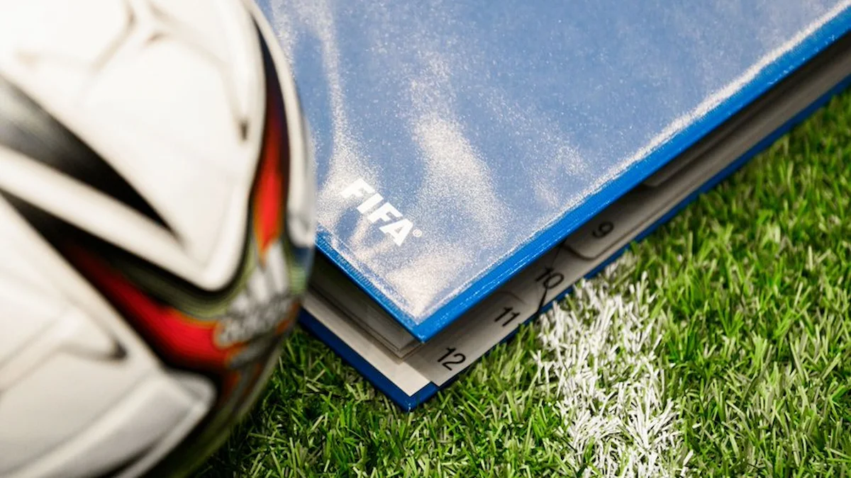 Balón de fútbol junto a una carpeta azul con el logo de la FIFA sobre el césped