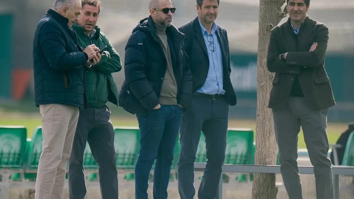 Cinco directivos conversan junto al césped durante un entrenamiento, con las gradas verdes al fondo