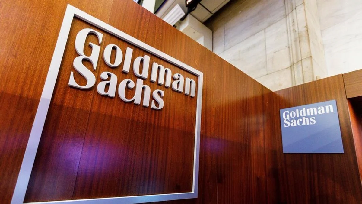 Logotipo de Goldman Sachs en letras plateadas sobre un panel de madera, con un cartel azul del banco al fondo